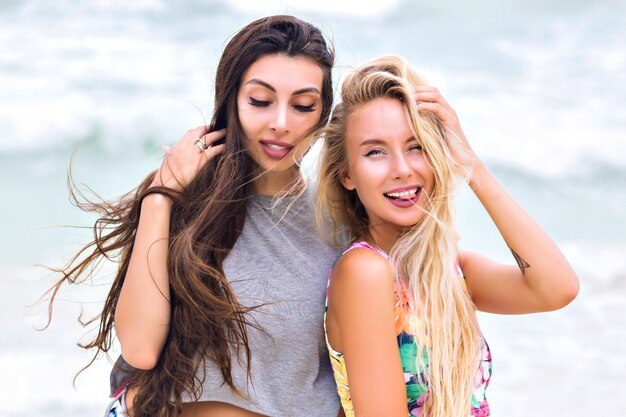 Retrato al aire libre de una chica guapa con cabello castaño oscuro posando con los ojos cerrados sobre fondo borroso del mar. Atractiva joven rubia con tatuaje de brazo divirtiéndose con un amigo durante las vacaciones de verano.