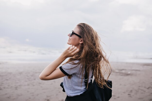 Retrato al aire libre de una chica increíble con cabello largo y oscuro que expresa felicidad durante el paseo por la playa. Modelo de mujer inspirada en camiseta gris pasar tiempo cerca del mar en un día nublado.