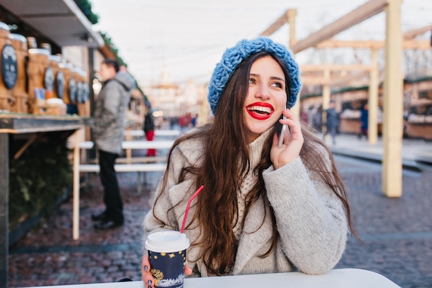 Foto gratuita retrato al aire libre de chica morena emocionada en abrigo de lana disfrutando de fin de semana de invierno en un día cálido. foto de dama caucásica de pelo largo con lindo sombrero azul posando en la calle borrosa