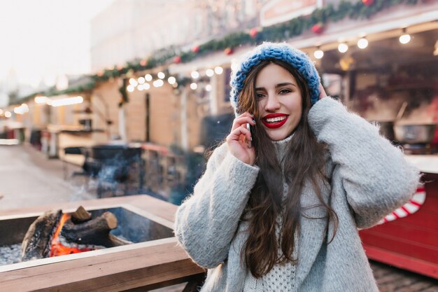 Retrato al aire libre de chica morena emocionada en abrigo de lana disfrutando de fin de semana de invierno en un día cálido. Foto de dama caucásica de pelo largo con lindo sombrero azul posando en la calle borrosa