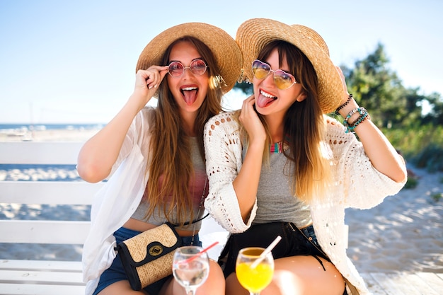 Foto gratuita retrato al aire libre de las chicas hipster divertidas felices de la compañía volviéndose locas en el café de la playa, bebiendo deliciosos cócteles riendo y sonriendo, trajes de verano boho brillante vintage, relaciones y diversión.