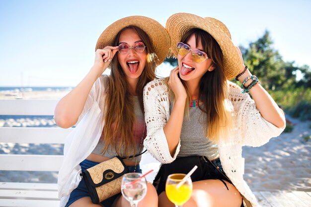 Retrato al aire libre de las chicas hipster divertidas felices de la compañía volviéndose locas en el café de la playa, bebiendo deliciosos cócteles riendo y sonriendo, trajes de verano boho brillante vintage, relaciones y diversión.