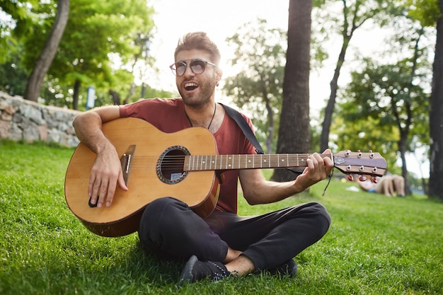 Retrato al aire libre de chico guapo hipster despreocupado sentado en el césped en el parque y tocando la guitarra