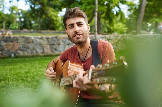 Retrato al aire libre de chico guapo romántico sentado en el césped en el parque y tocando la guitarra