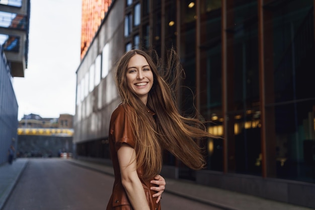 Retrato al aire libre en la cintura de una mujer carismática hermosa que se divierte sintiéndose feliz riendo mientras vuelve a sonreír con la cámara caminando por la calle vacía de la ciudad cerca de los edificios comerciales