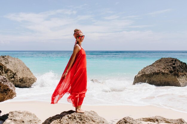 Retrato al aire libre de cuerpo entero de una chica inspirada sonriente posando en una gran piedra Espectacular dama viste un largo vestido rojo mirando a la cámara mientras está de pie frente al mar azul