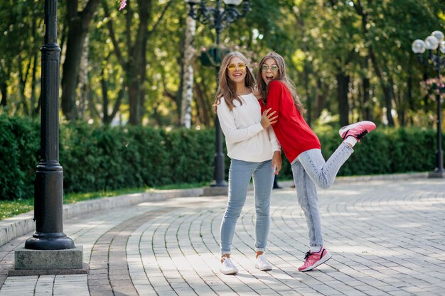 Retrato al aire libre de cuerpo entero de dos niñas felices muy amables que se divierten y caminan juntas después de estudiar en la ciudad, día soleado, buenas emociones verdaderas, humor divertido