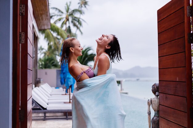 Retrato al aire libre de dos mujeres felices en bikini con toallas de playa de vacaciones fuera de la villa junto a la piscina en un día lluvioso