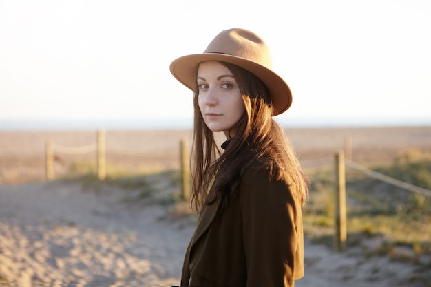 Retrato al aire libre de una elegante joven europea con sombrero de moda y abrigo negro mirando con una sutil sonrisa mientras pasea por el mar, sueña y admira la puesta de sol