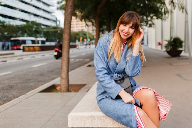 Retrato al aire libre de elegante mujer muy feliz en abrigo azul sentado en un banco en la calle