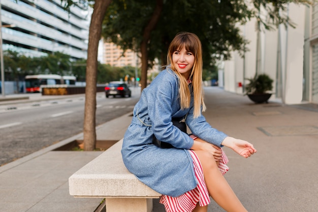 Retrato al aire libre de elegante mujer muy feliz en abrigo azul sentado en un banco en la calle