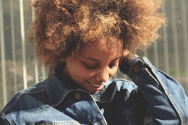 Foto gratuita retrato al aire libre de una elegante niña africana con corte de pelo afro mirando hacia abajo con expresión alegre y despreocupada.