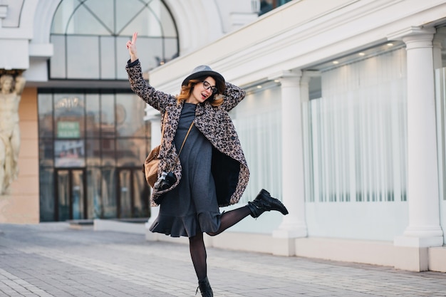 Retrato al aire libre de elegante señorita con mochila marrón con abrigo y sombrero. Mujer atractiva con el pelo rizado hablando saltando y divirtiéndose.