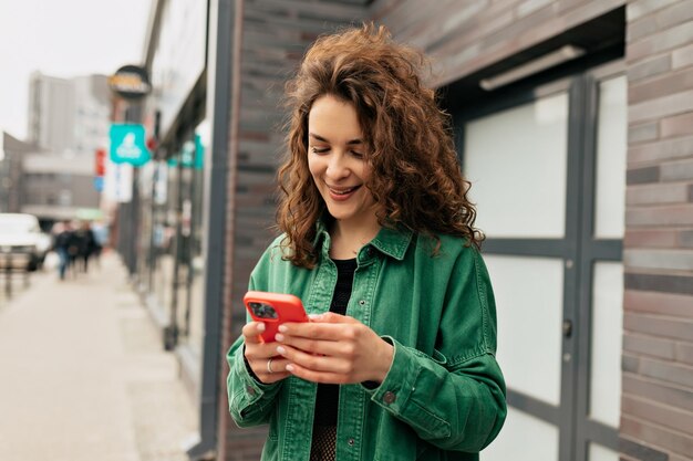 Retrato al aire libre de una encantadora chica elegante con rizos con camisa verde usando un teléfono inteligente con una sonrisa Una joven caucásica despreocupada está usando un teléfono inteligente moderno parado al aire libre