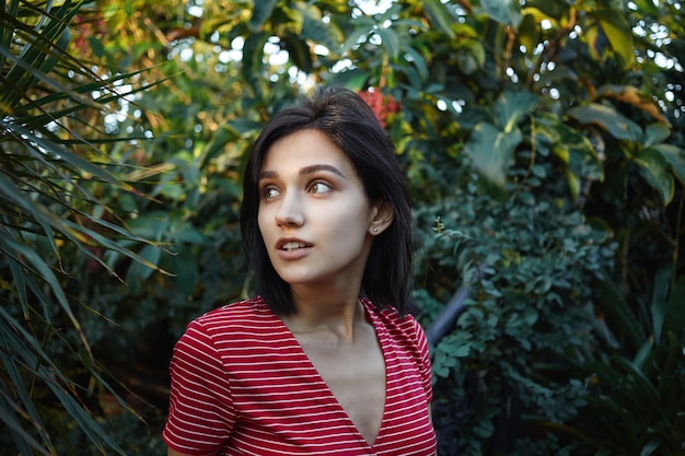 Retrato al aire libre de una encantadora y misteriosa chica veinteañera que se relaja entre plantas verdes frescas mientras pasa las vacaciones en una isla tropical, camina en la jungla, estudia la flora y fauna local