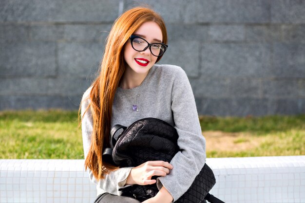 Retrato al aire libre de estilo de vida de la muchacha bonita de jengibre con cristal transparente y suéter gris de cachemira, posando en el parque, estilo otoño invierno Sosteniendo una mochila.