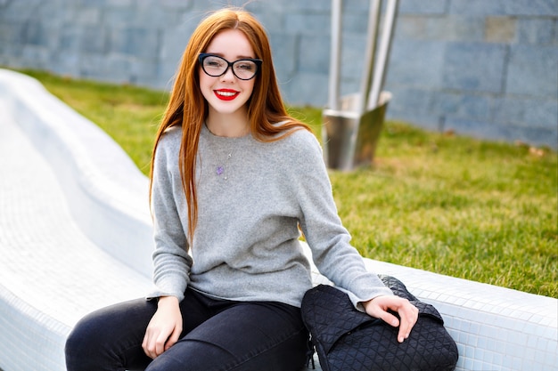 Retrato al aire libre de estilo de vida de la muchacha bonita de jengibre con cristal transparente y suéter gris de cachemira, posando en el parque, estilo otoño invierno
