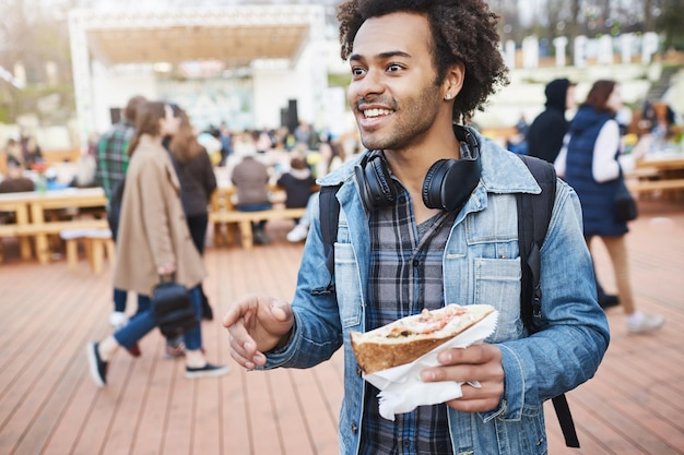 Retrato al aire libre de un estudiante afroamericano positivo sosteniendo un sándwich mientras gesticula y habla con un amigo, caminando en el festival, esperando el concierto de su banda favorita.