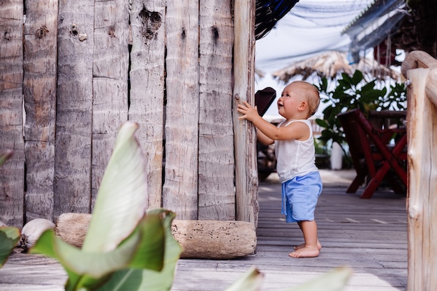 Retrato al aire libre de feliz niño de nueve meses en camisa azul corta y blanca se encuentra junto a una pared de madera y una sonrisa