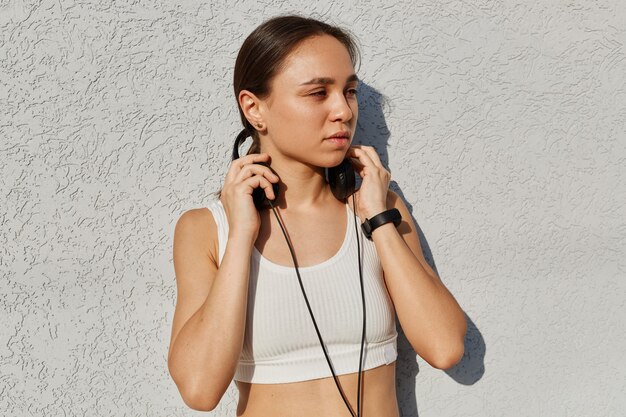 Retrato al aire libre de una hermosa mujer adulta joven con top blanco, escuchando música durante el entrenamiento, manteniendo las manos en los auriculares, mirando a otro lado con expresión facial pensativa.