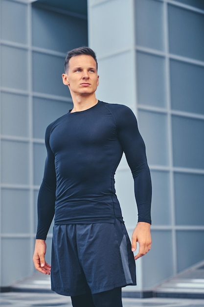 Foto gratuita retrato al aire libre de un hombre atlético y atractivo vestido con ropa deportiva negra sobre un edificio moderno.