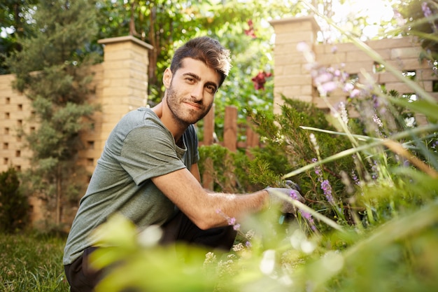 Retrato al aire libre de hombre caucásico barbudo atractivo joven en camisa azul y pantalones deportivos sonriendo, sentado en la hierba, mirando a puerta cerrada con expresión de cara feliz, trabajando en el jardín.