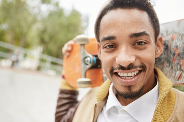 Retrato al aire libre de un hombre guapo con ojos oscuros, barba y bigote, mantiene la patineta detrás de la cabeza