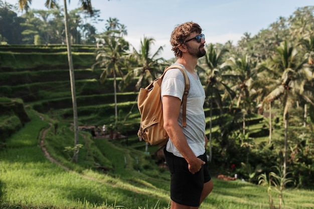 Retrato al aire libre del hombre de viaje guapo con mochila caminando en terraza de subida en Bali.