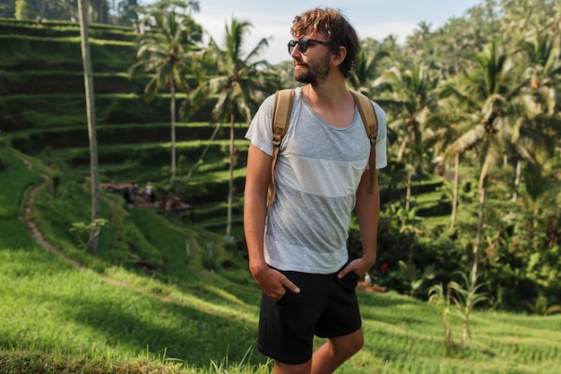 Retrato al aire libre del hombre de viaje guapo con mochila caminando en terraza de subida en Bali.