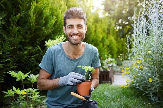 Retrato al aire libre de joven guapo caucásico barbudo con camisa azul y guantes sonriendo en la cámara, sosteniendo la olla con flor en las manos trabajando en el jardín.