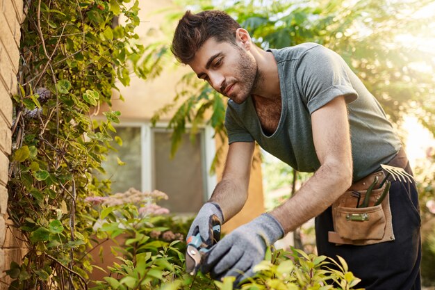 Retrato al aire libre de joven hispano barbudo atractivo en camiseta azul y guantes trabajando en el jardín con herramientas, cortando hojas, regando las plantas. La vida rural