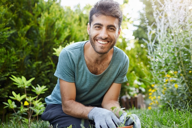 Retrato al aire libre de joven jardinero masculino caucásico barbudo atractivo en camiseta azul sonriendo en la cámara, plantando semillas en el jardín, regando plantas de cerca.