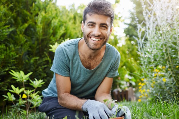 Retrato al aire libre de joven jardinero masculino caucásico barbudo atractivo en camiseta azul sonriendo en la cámara, plantando semillas en el jardín, regando plantas de cerca.