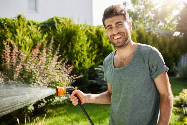 Retrato al aire libre de joven jardinero masculino caucásico guapo sonriendo regando las plantas, pasando el verano en casa de campo.