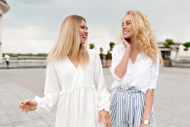 Retrato al aire libre de lindas damas que se divierten afuera relajándose en la ciudad de verano mientras caminan por la calle y se ríen en un día nublado de verano. Dos chicas con estilo afuera