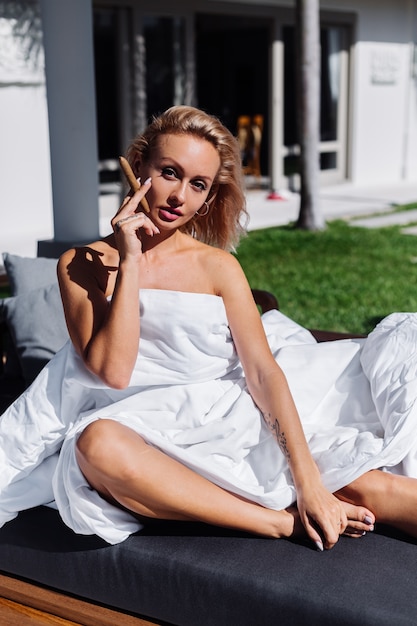 Retrato al aire libre de moda de mujer desnuda sentada en el sofá cubriéndose con una manta con cigarro