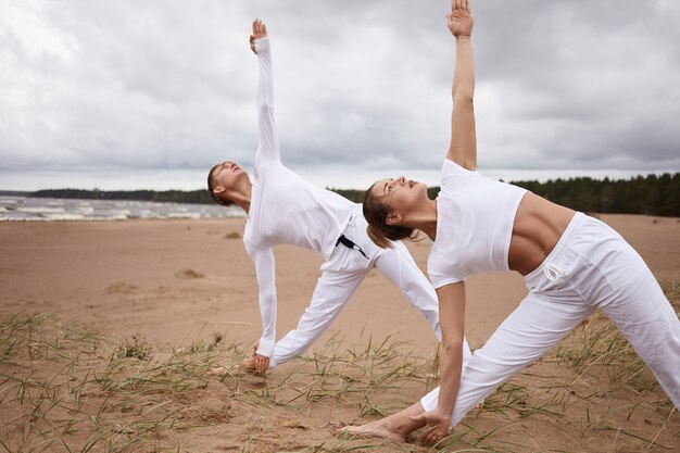 Retrato al aire libre de una mujer atractiva y un hombre joven con cuerpos atléticos, ambos vestidos con trajes blancos, practicando yoga en la playa durante el retiro, haciendo Utthita Trikonasana o postura de triángulo extendido