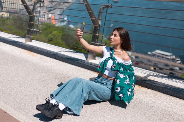 Retrato al aire libre de mujer en camisa verde casual en día soleado se encuentra en el puente mirando en la pantalla del teléfono tomar selfie hacer videollamada