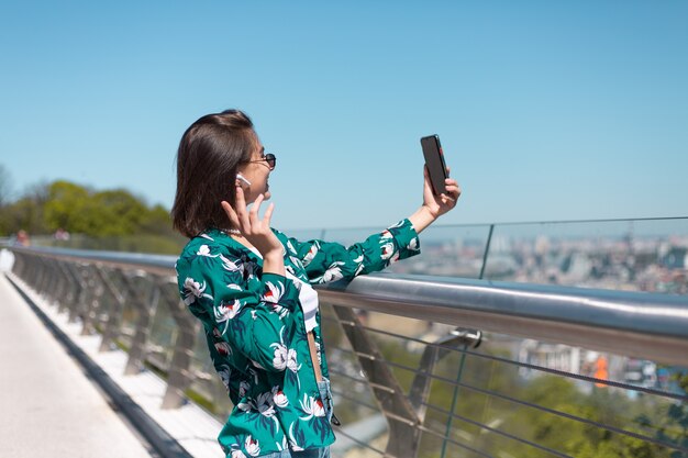 Retrato al aire libre de mujer con camisa verde casual en día soleado se encuentra en el puente mirando en la pantalla del teléfono tomar selfie hacer videollamadas auriculares inalámbricos bluetooth en los oídos