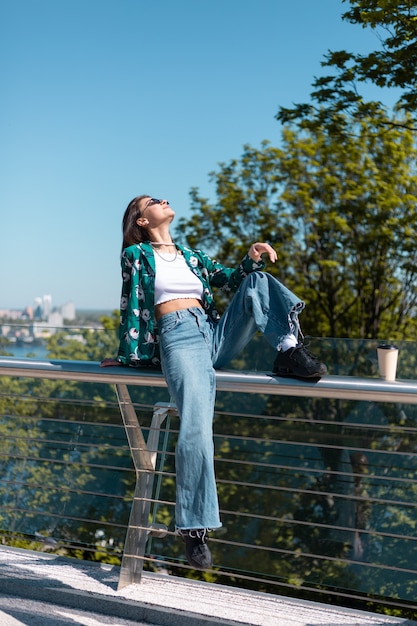 Retrato al aire libre de mujer con camisa verde casual y jeans en un día soleado en el puente con una vista increíble de la ciudad en la mañana