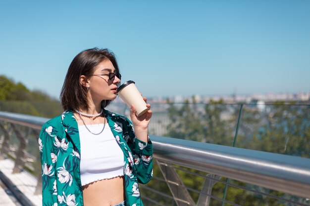 Retrato al aire libre de mujer con camisa verde con una taza de café disfrutando del sol, se encuentra en el puente con una vista increíble de la ciudad en la mañana