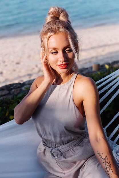 Retrato al aire libre de mujer caucásica en mono clásico con lápiz labial rojo en una hamaca de vacaciones fuera del hotel villa, junto al mar