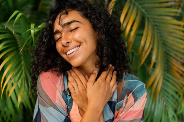 Retrato al aire libre de una mujer encantadora y dichosa con pelos rizados posando sobre árboles tropicales y hojas de palma