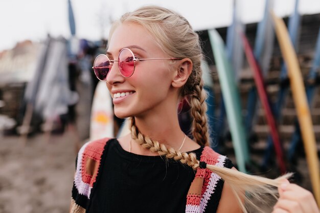 Retrato al aire libre de mujer rubia feliz con gafas de sol rosas mirando a otro lado sobre fondo borroso.