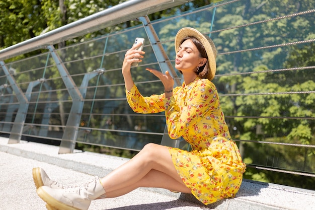 Retrato al aire libre de mujer en vestido amarillo de verano sentado en el puente tomar selfie en teléfono móvil