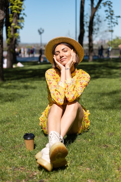 Retrato al aire libre de mujer en vestido amarillo de verano y sombrero sentado sobre el césped en el parque