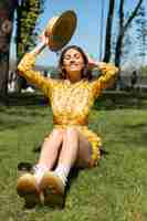 Foto gratuita retrato al aire libre de mujer en vestido amarillo de verano y sombrero sentado sobre el césped en el parque