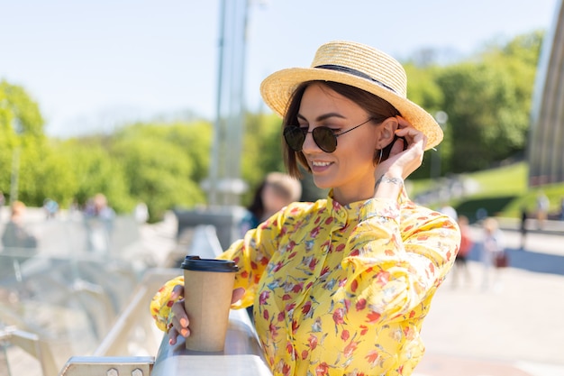 Retrato al aire libre de mujer con vestido amarillo de verano y sombrero con una taza de café disfrutando del sol, se encuentra en el puente con una vista increíble de la ciudad