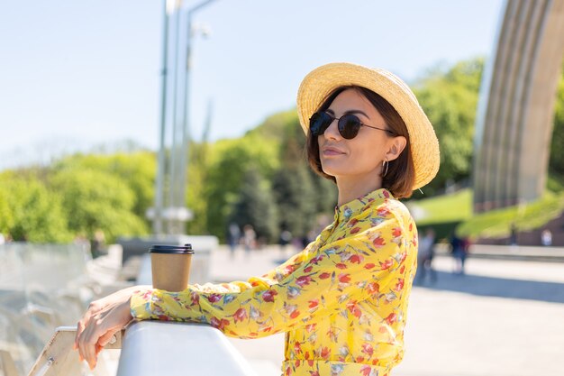 Retrato al aire libre de mujer con vestido amarillo de verano y sombrero con una taza de café disfrutando del sol, se encuentra en el puente con una vista increíble de la ciudad