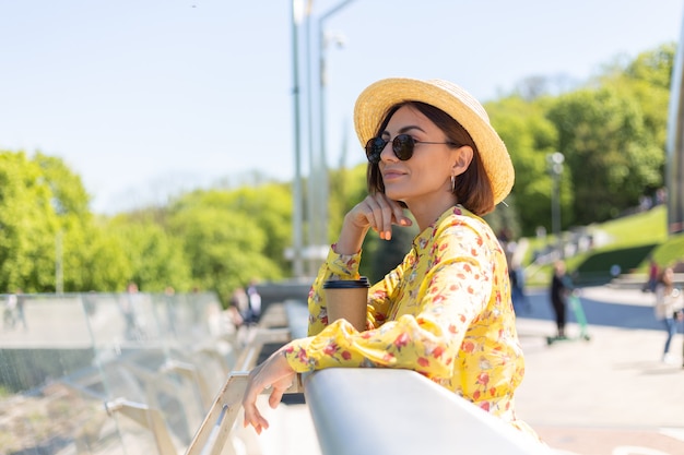 Retrato al aire libre de mujer con vestido amarillo de verano y sombrero con una taza de café disfrutando del sol, se encuentra en el puente con una vista increíble de la ciudad